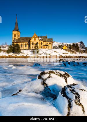 The wooden yellow Vagan Church on the beach on the Lofoten islands in Norway in winter with frozen river and mountains in morning light Stock Photo