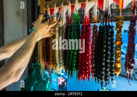 Churchkhela (Georgian ჩურჩხელა) in white, yellow, green, red, brown and black hangs in strands at a stall in Tbilisi, Georgia. A hand uses scissors to cut one strand from the rail. The person wears a turquoise bracelet on the wrist. To make this traditional Georgian sweet, walnuts or hazelnuts are threaded onto a string. The strands are dipped several times into pelamushi, grape juice boiled down and thickened with flour, and then dried. This gives them the shape of a candle. Churchkhela is a classic provision for hiking in the Caucasus. The traditional method of making it in Kakheti has been  Stock Photo