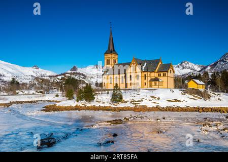 The wooden yellow Vagan Church on the beach on the Lofoten islands in Norway in winter with frozen river and mountains in morning light Stock Photo