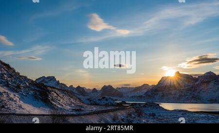 Scenic view over sun setting behind snow-clad mountain with road and lake on the Lofoten islands archipelago in Norway Stock Photo