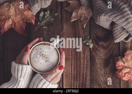 Female hands holding hot coffee cappucino, blanket and autumn leaves on old wooden background. Vintage tonning. autumn relax concept Stock Photo