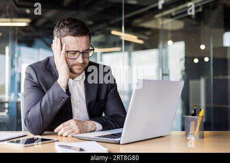 Problems at work. Upset young male office worker, businessman sitting at a desk in front of a laptop and thoughtfully leaning his head on his hand. Stock Photo