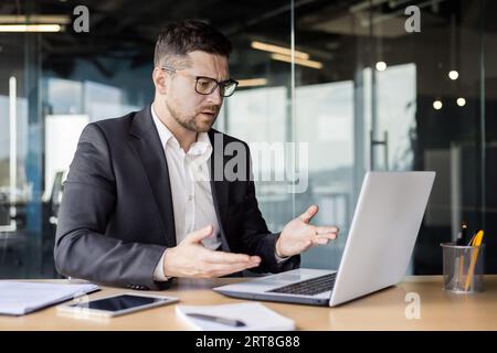 A worried young man businessman sits in the office in front of a laptop and looks sadly at the camera, spreading his hands. Stock Photo