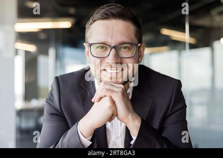 Close-up photo of a young smiling man, businessman, director, sitting in the office in a suit, resting his head on his hands and looking at the camera with a smile. Stock Photo