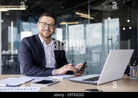 Portrait of a young businessman, accountant, financial analyst working in the office with a calculator and documents, sitting at a desk with a laptop and smiling at the camera. Stock Photo