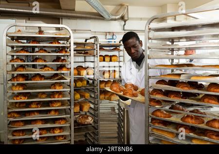 African baker arranging trays with bakery products on trolley Stock Photo