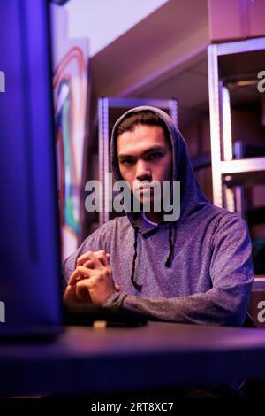 Serious hacker developing malicious software and looking at camera. Young asian criminal in hood doing online illegal activities and programming malware on computer portrait Stock Photo