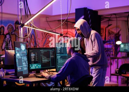 Hackers coding malicious software on computer together while working in abandoned warehouse. Internet thieves in hoods planning phishing attack and programming malware in dark room Stock Photo