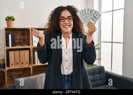 Young hispanic woman holding dollars screaming proud, celebrating victory and success very excited with raised arm Stock Photo