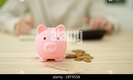 Young hispanic man writing on notebook using calculator with piggy bank and coins on table on table Stock Photo