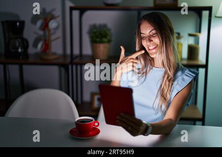 Young hispanic woman using touchpad sitting on the table at night smiling cheerful showing and pointing with fingers teeth and mouth. dental health co Stock Photo