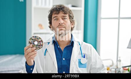 Young hispanic man nutritionist holding doughnut with serious face at clinic Stock Photo