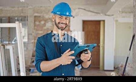 Young hispanic man architect wearing hardhat smiling using tablet at construction site Stock Photo