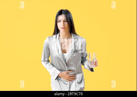 Young woman with glass of water suffering from menstrual cramps on yellow background Stock Photo
