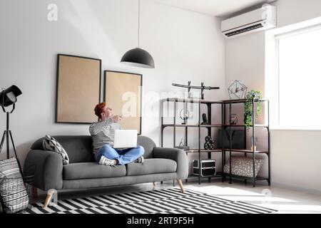 Young man with laptop switching on air conditioner at home Stock Photo