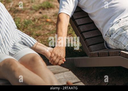 A couple of cute elderly people lie on sunbeds holding hands, basking in the sun. Close-up of hands Stock Photo