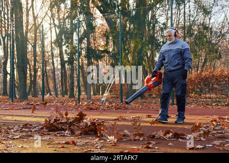 Senior man in overalls, headphones and gloves cleaning fenced city park area from dry fall leaves. Side view of professional janitor using hands blower during working process outside. Work concept. Stock Photo