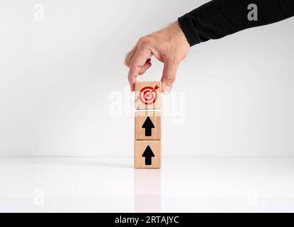 Goal achievement and business objectives. Goal setting. Aspiring for higher goals. Hand stacking the wooden cubes with arrow and target goal symbols. Stock Photo