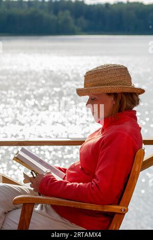 A woman sitting reading in a chair wearing a straw hat on a jetty Stock Photo