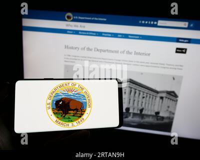 Person holding cellphone with seal of United States Department of the Interior (DOI) on screen in front of webpage. Focus on phone display. Stock Photo