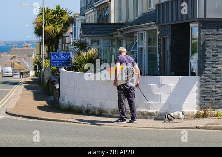 A mature man carrying a surfboard and his pet terrier walking along a street in Newquay Town Centre in Cornwall in the UK. Stock Photo