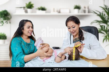 Female pediatrician examines 6 months baby girl and measures the growth. Doctor using measurement tape to check baby head size while mother holding he Stock Photo