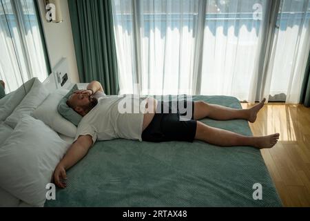 Lazy exhausted overweight man sleeping in bed feeling bad on vacation in hotel room. Stock Photo