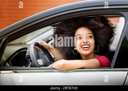 Smiling young Moroccan female with Afro hairstyle looking away while sitting in driver seat of parked car in daylight with hand on steering wheel and Stock Photo