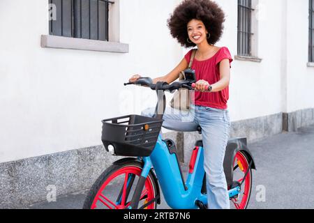 Enjoying young Moroccan woman with Afro hairstyle smiling and looking at camera while riding electric bicycle and standing with foot on street Stock Photo