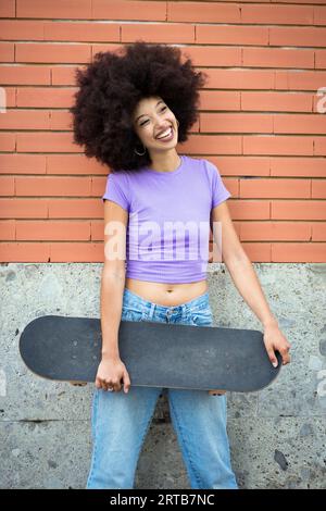Positive African American female with Afro hairstyle and in casual clothes looking at camera while leaning on brick wall with skateboard Stock Photo