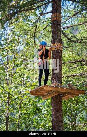 Low angle of teenage girl in helmet looking away while standing on wooden board on tree trunk in green forest and holding belay cable attached to rope Stock Photo