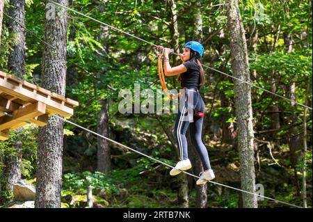 Side view of happy young female in protective helmet looking away while walking on rope with safety cables attached to top wire and enjoying adventure Stock Photo