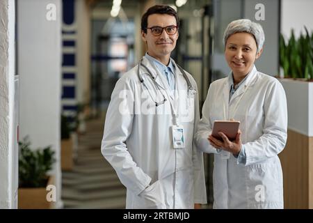 Two confident experienced general practitioners in lab coats looking at camera while standing in corridor of clinics and having discussion Stock Photo
