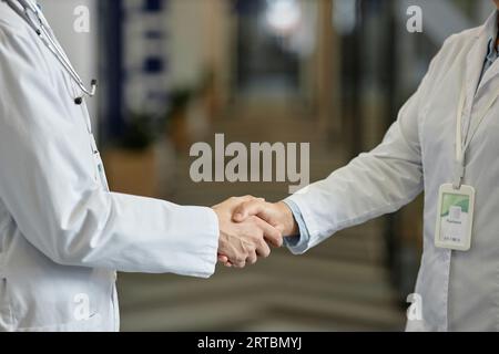 Close-up of hands of two general practitioners in lab coats standing in front of camera in corridor of modern hospital during handshake Stock Photo