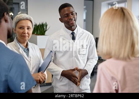 Two confident male and female clinicians in lab coats talking to their assistants at meeting and discussing purchase of new medical equipment Stock Photo