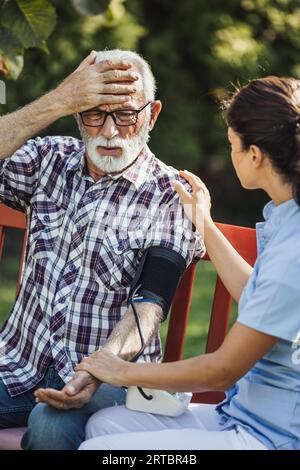 Senior man having headache and young woman caregiver checking his blood pressure Stock Photo