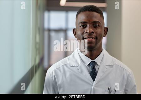 Happy young African American male general practitioner in lab coat and tie looking at camera while standing in corridor of modern clinics Stock Photo