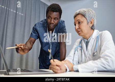 Young African American assistant of general practitioner pointing at screen of laptop during explanation of new methods of medical treatment Stock Photo