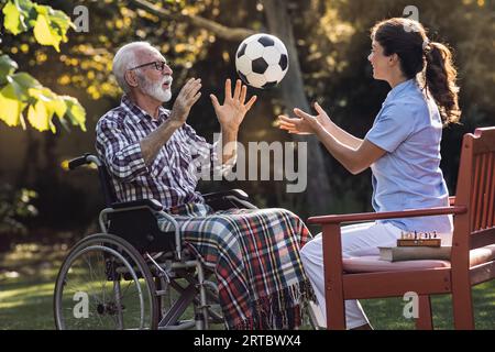 Senior man in wheelchair doing workout by throwing football to young woman caregiver in park Stock Photo