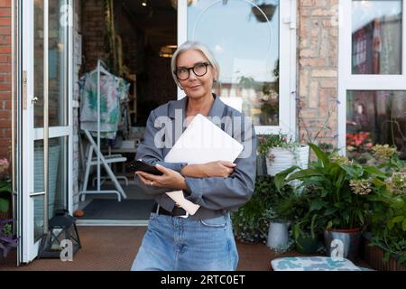 well-groomed senior woman with gray hair and glasses dressed in a gray jacket and jeans works remotely and walks along the street holding a laptop Stock Photo