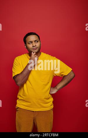 pensive indian man in bright casual attire standing with hand near chin on red background in studio Stock Photo