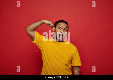 curious indian man in bright casual clothes looking away and touching ear on red background Stock Photo