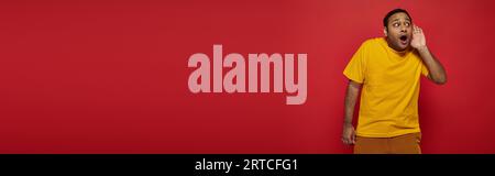 curious indian man in bright clothes looking away and listening on red background, banner Stock Photo