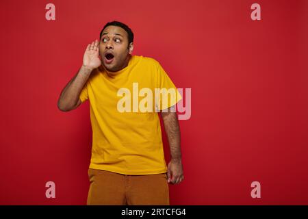 curious indian man in bright clothes looking away and listening on red backdrop, hand near ear Stock Photo