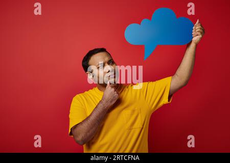 thoughtful indian man in yellow t-shirt holding blank thought bubble on red backdrop, pensive face Stock Photo