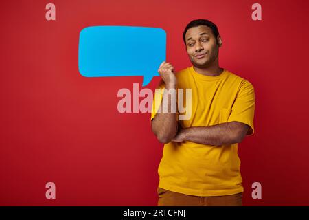 skeptical indian man in yellow t-shirt holding blank speech bubble on red background Stock Photo