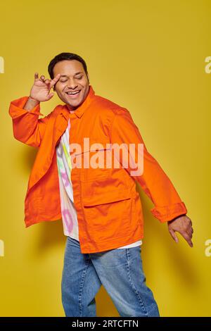 portrait of happy indian man in orange jacket and denim jacket dancing on yellow background Stock Photo