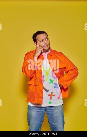 portrait of pensive indian man in orange jacket posing with hand on hip on yellow background Stock Photo