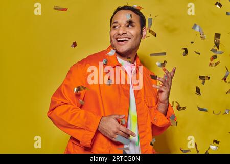 happy indian man in bright orange jacket smiling near falling confetti on yellow backdrop, party Stock Photo