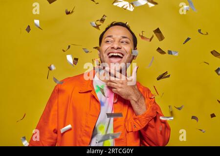 joyous indian man in bright orange jacket smiling near falling confetti on yellow backdrop, party Stock Photo